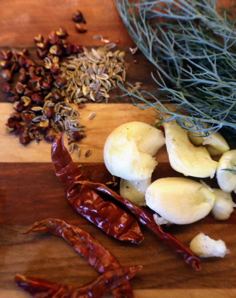 Pickle ingredients on wooden cutting board including Thai dried peppers, fennel seeds, smashed garlic cloves, and fresh dill